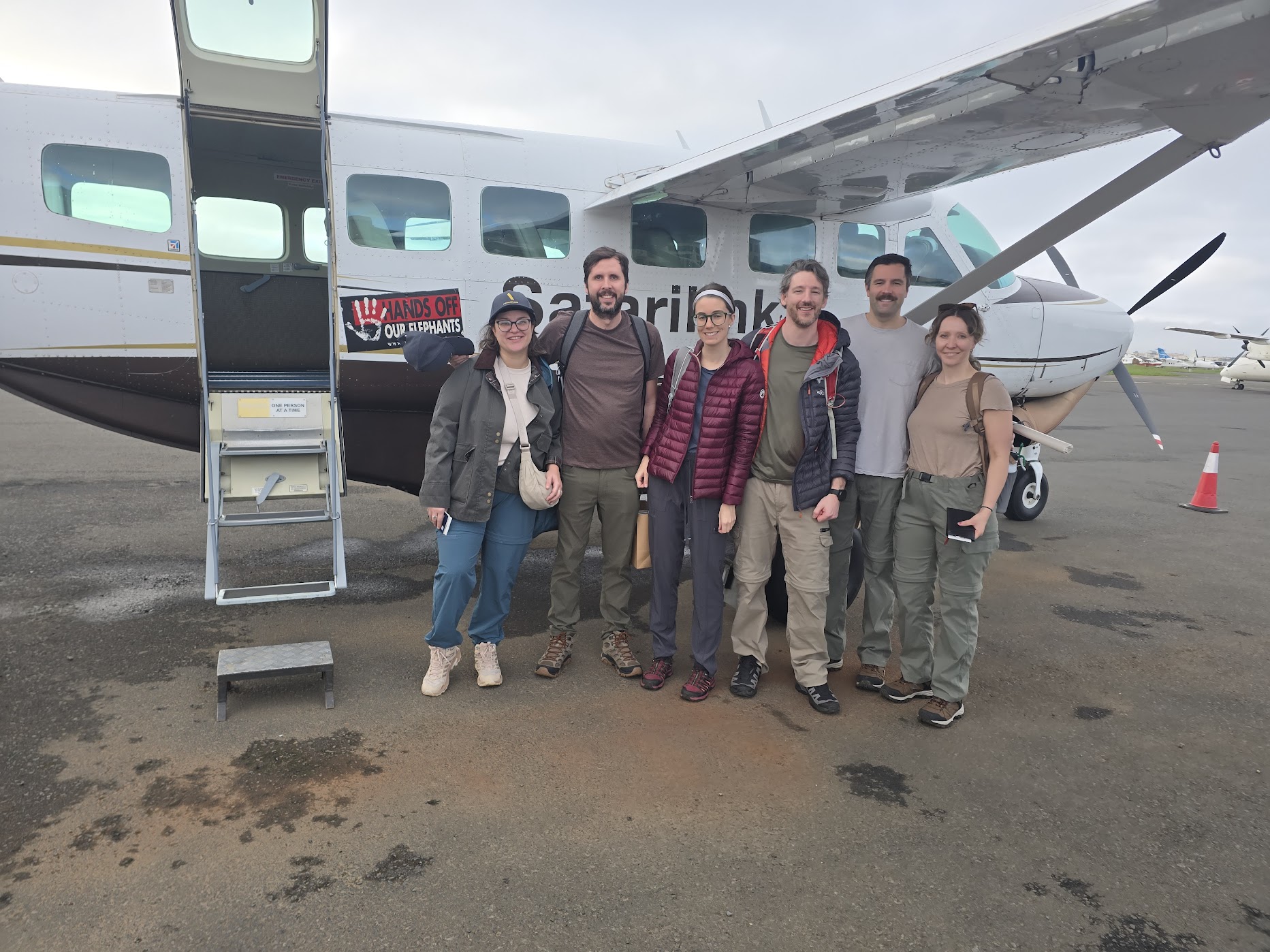 Six travellers in earth-toned safari clothing stand together beside a white Safarilink Cessna Caravan on the tarmac under an overcast Nairobi sky before the flight to Ol Pejeta.