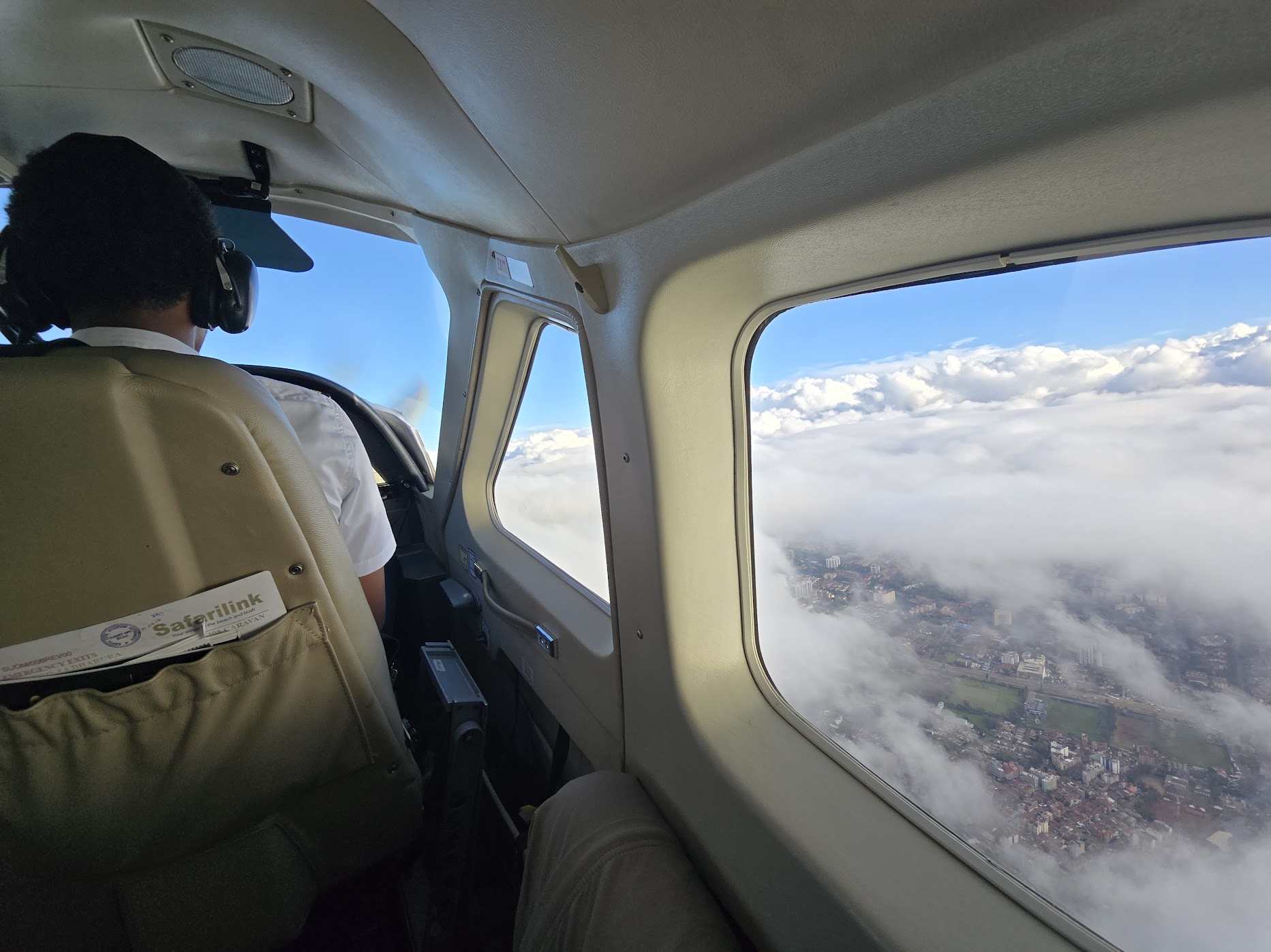 View forward inside the Safarilink Caravan cabin: the pilot in a white shirt and headset at the controls, with Nairobi dissolving beneath a broken layer of cloud through the oval window.