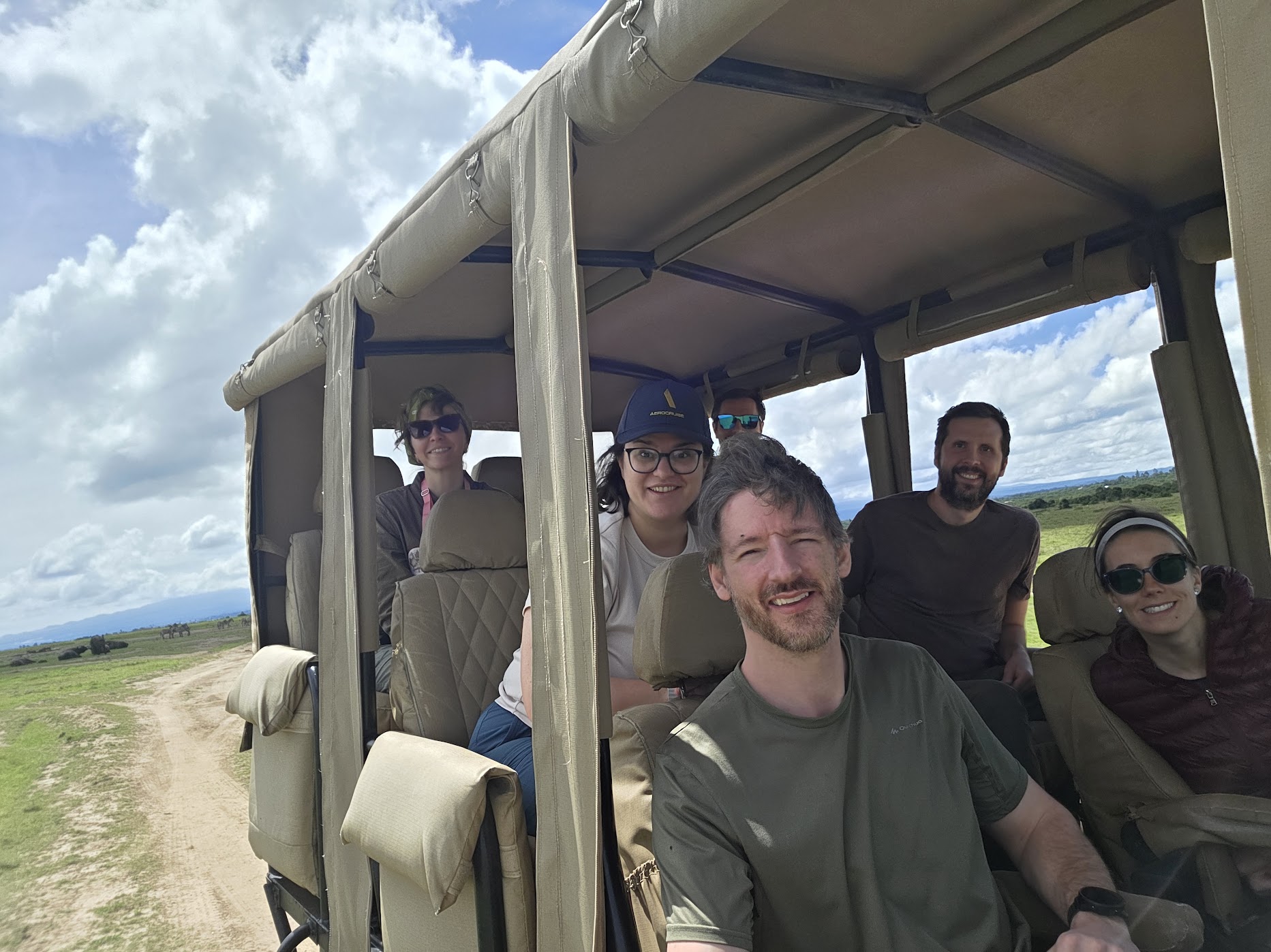 Six safari guests fill a canvas-sided open game vehicle on the Ol Pejeta savanna, rolled side flaps up, lush grassland and building cloud behind them.