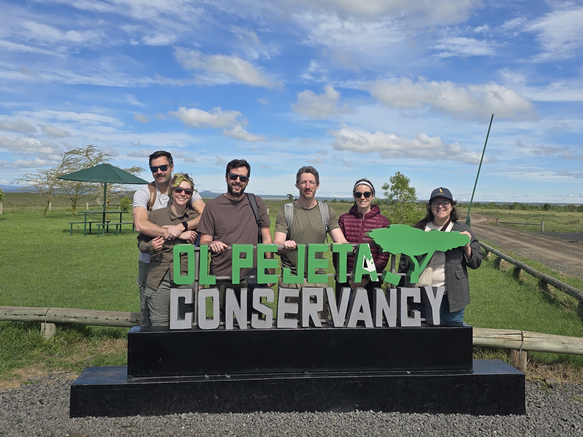 Six travellers pose behind the green-and-white Ol Pejeta Conservancy sign on a black plinth, wide grassland and blue sky scattered with high cloud behind them.
