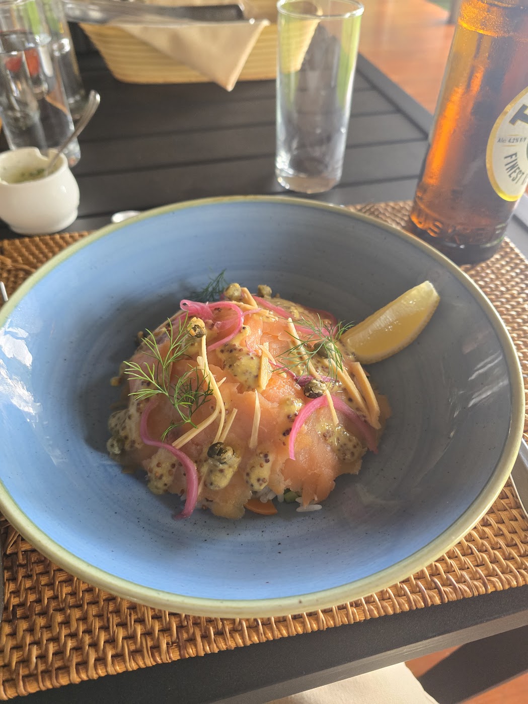 A plated course of cured salmon in a matte blue-grey ceramic bowl, draped in whole-grain mustard with pickled red onion ribbons, fresh dill and a lemon wedge, on a woven rattan placemat in warm outdoor light.