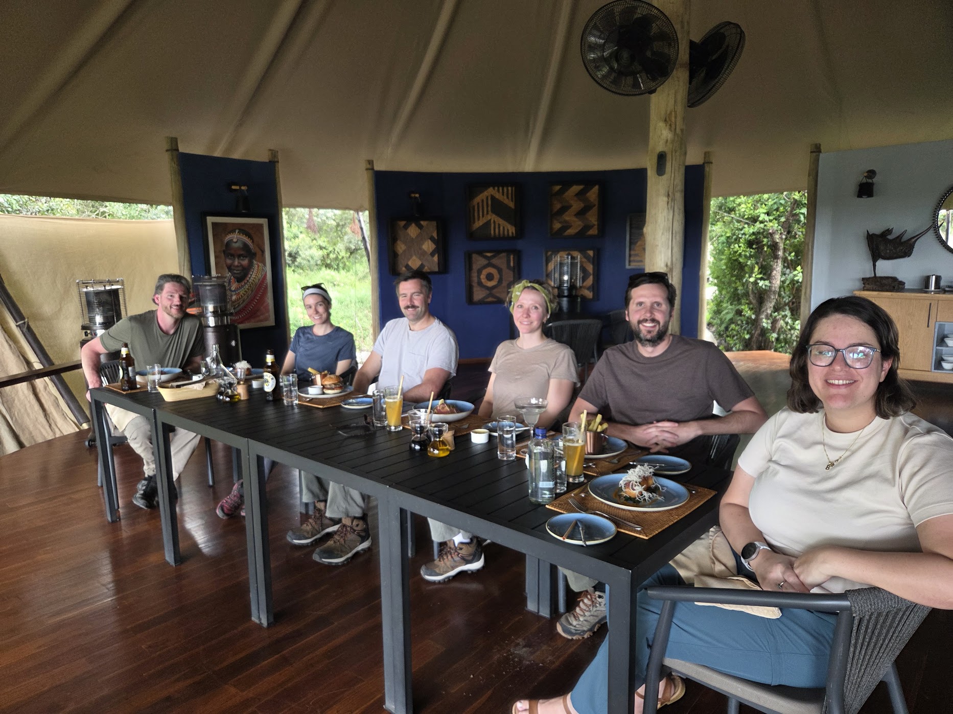Six travellers seated along a dark dining table inside a canvas safari tent at Ol Pejeta, warm interior light, a cobalt-blue feature wall hung with geometric woven art and a portrait behind them.
