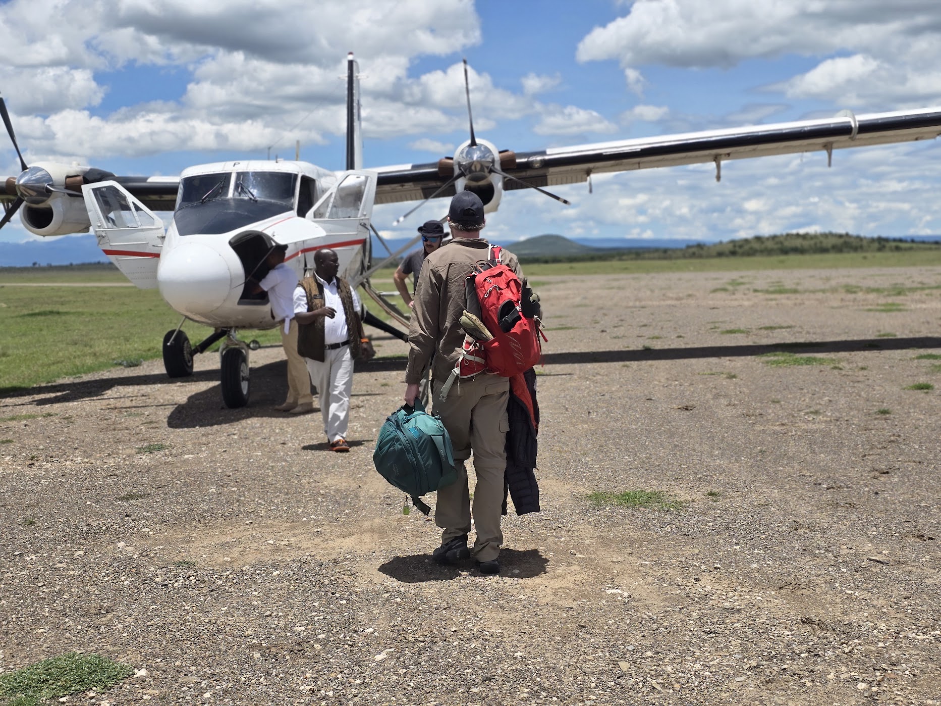 A traveller with a red backpack and teal duffel walks across a dirt airstrip toward a white twin-engine bush plane at Ol Pejeta, a greeter in white at the aircraft door under a bright mid-morning cloudscape.
