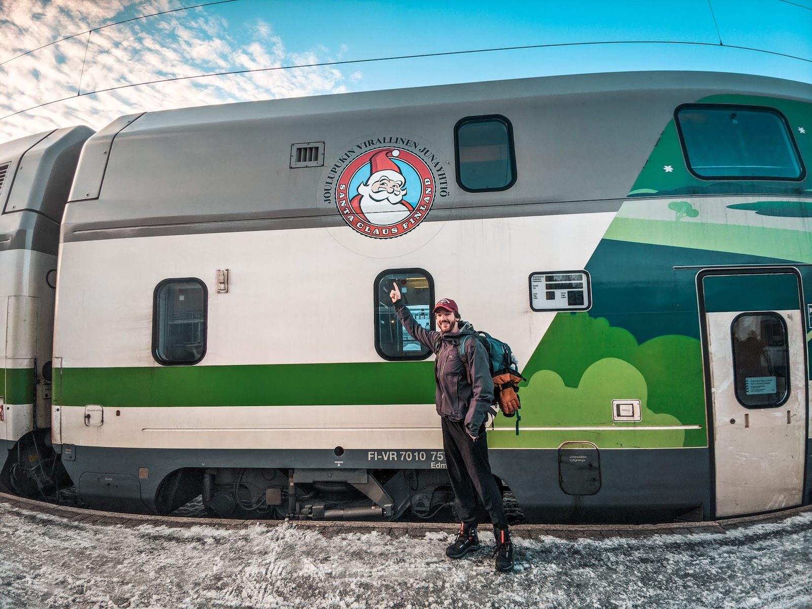 A traveller in a dark jacket points up at the Santa Claus Finland reindeer logo on the side of a green and white double-decker VR train on a snowy platform in Rovaniemi.
