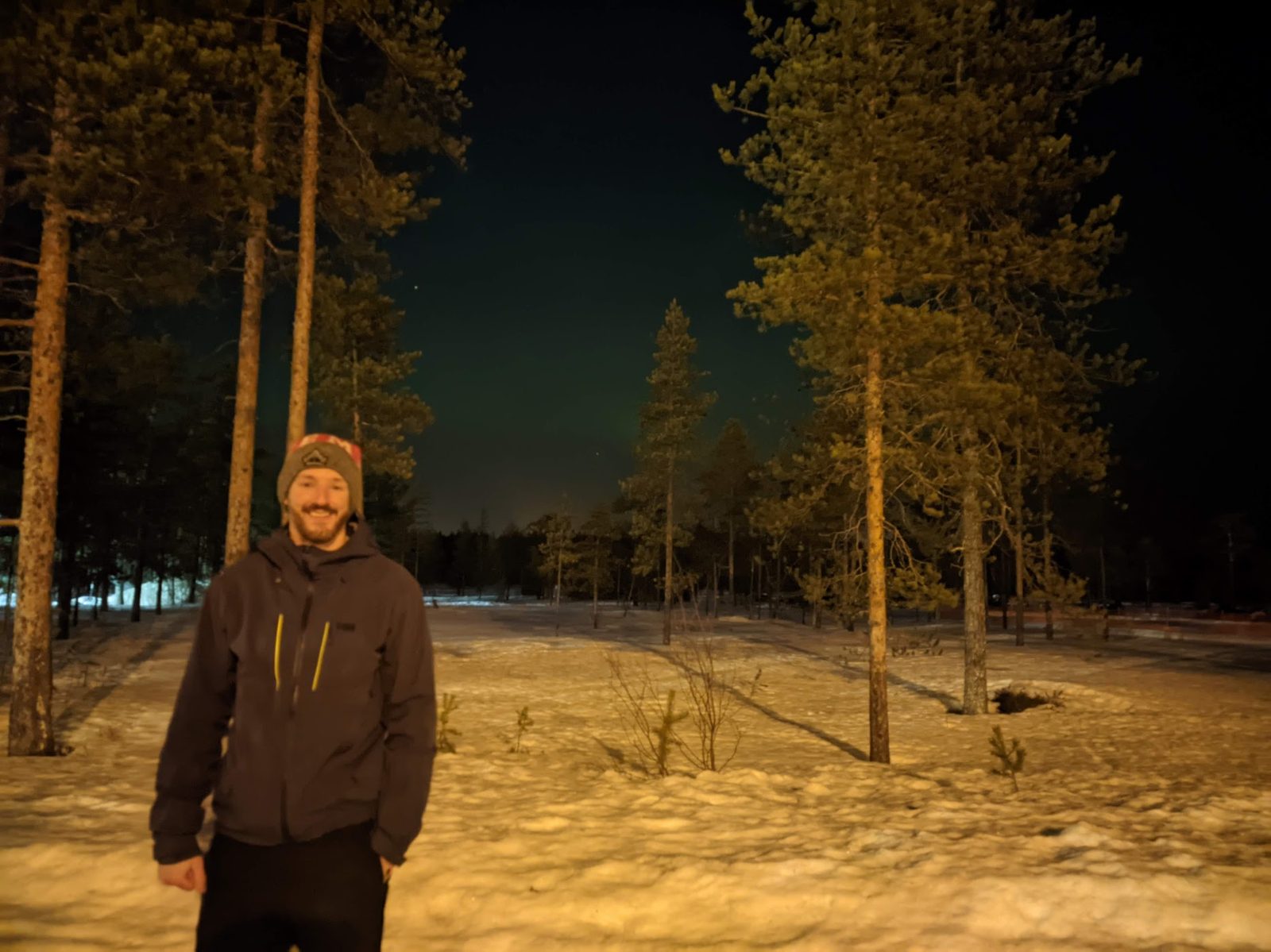 A man in a dark jacket and beanie stands among tall pines on snow at night, a faint green band of aurora glowing low in the sky behind the treeline.