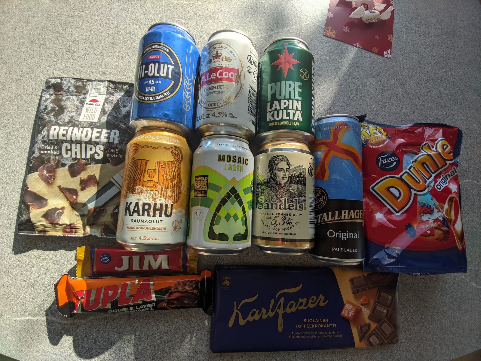 A spread of Finnish canned beers including Lapin Kulta, Karhu and Sandels laid out beside Fazer chocolate bars and a bag of reindeer chips on a grey table in sunlight.
