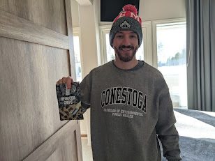 A smiling man in a grey sweater holds up a dark foil bag of Polarica dried and smoked reindeer chips in a hotel room.
