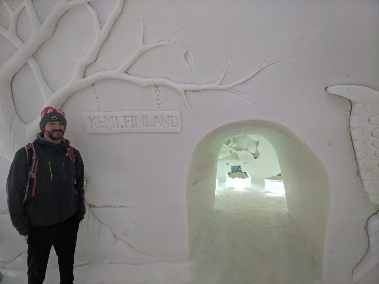 A man in a beanie and backpack stands inside the Kemi SnowCastle beside a carved snow relief of a bare tree and a sign reading KEMI.FINLAND, an arched snow tunnel leading deeper inside.