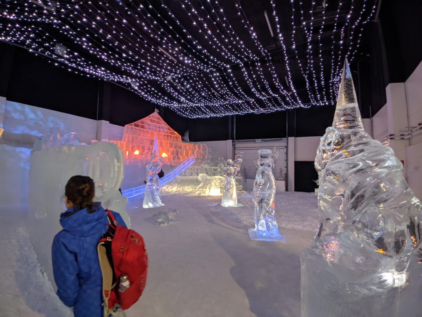 A child in a blue coat looks at a hall of backlit ice sculptures under a canopy of fairy lights inside the Kemi SnowCastle, a glowing orange circus tent carved in ice at the rear.