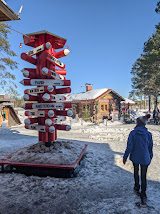 A man in a beanie stands beside the tall red Arctic Circle marker post at Santa Claus Village under a bright blue winter sky.