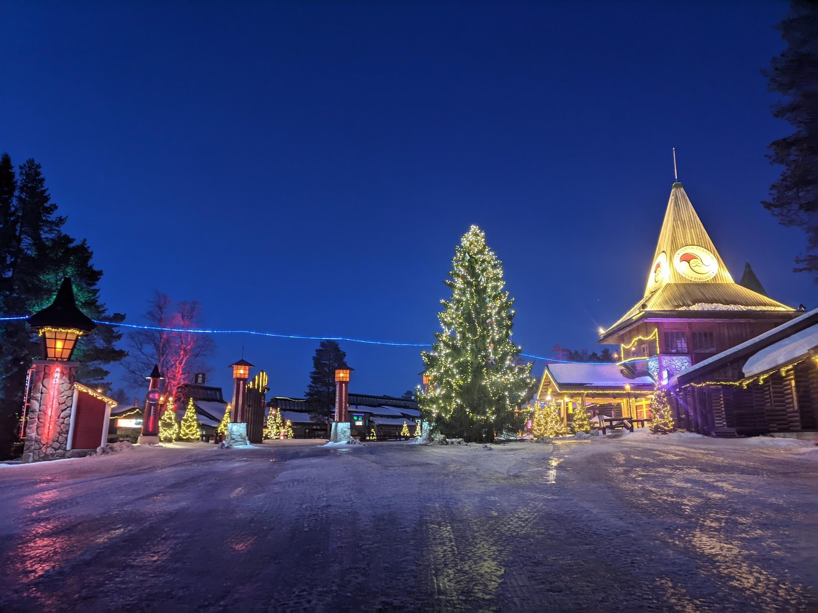 Santa Claus Village at blue hour: a fully lit Christmas tree at centre, the illuminated Arctic Circle tower at right, snow underfoot and a deep blue sky overhead.