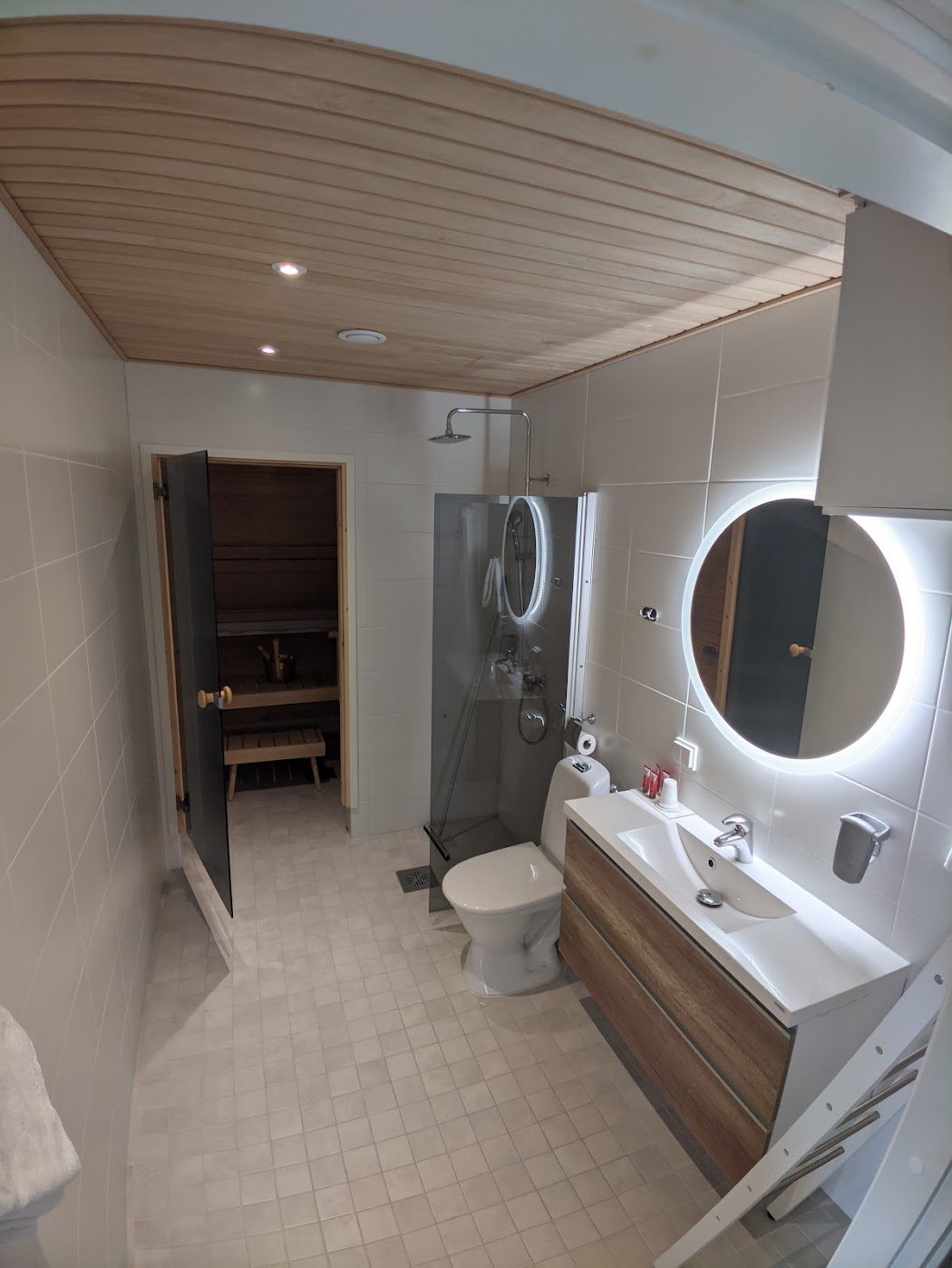 A clean Nordic hotel bathroom with a pale wood-slatted ceiling, a round backlit mirror, a glass walk-in shower and an open door to a small private wood-lined sauna.