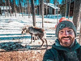 A smiling man takes a selfie crouched beside a reindeer with felted antlers in a snowy clearing fringed by pine and a red cabin.
