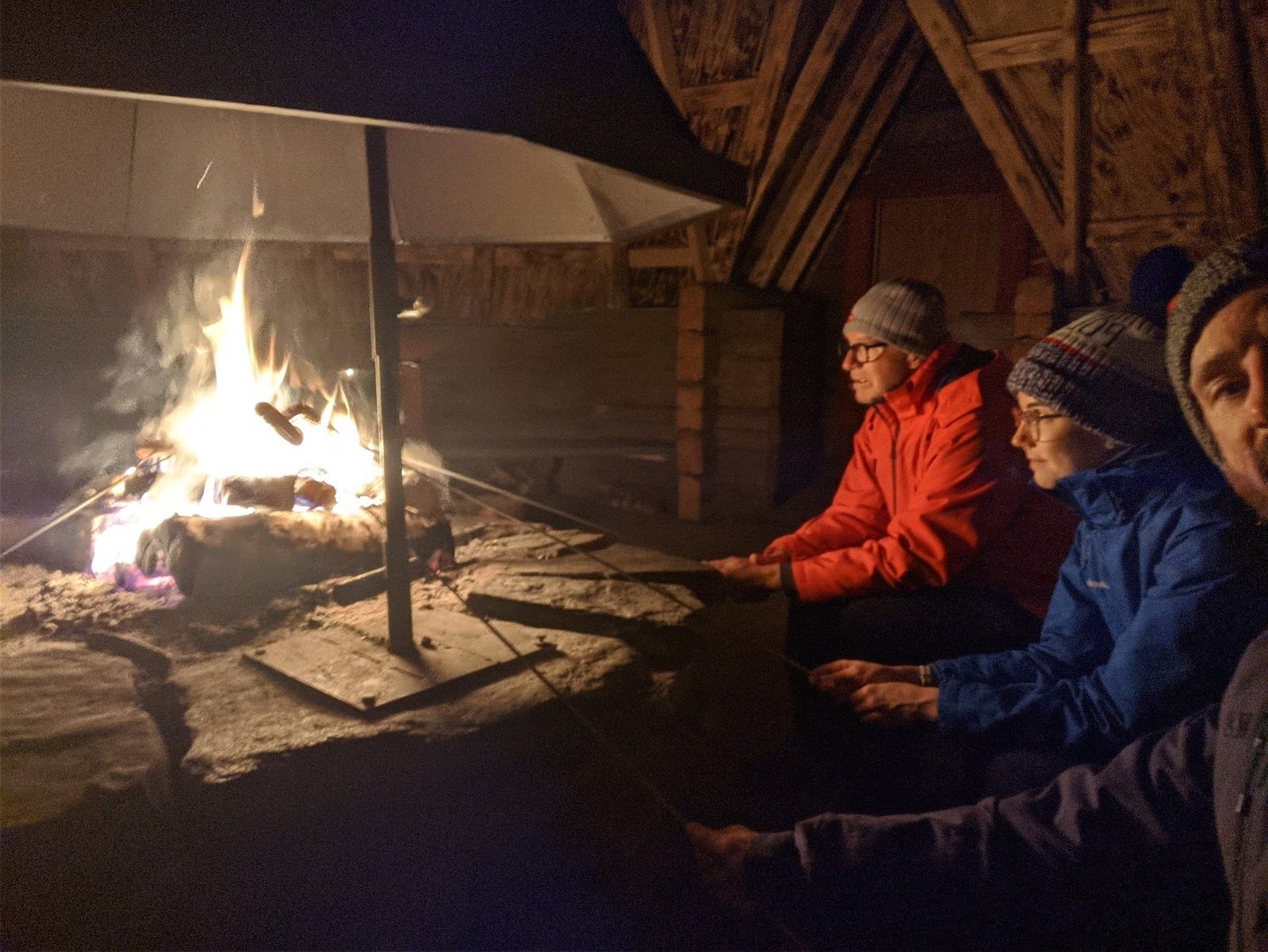 Three people in winter jackets sit on reindeer hides around an open fire under a wooden lavvu shelter at night, hands stretched toward the flames.